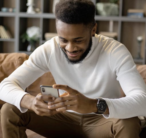 Man in a white ribbed long-sleeve shirt and tan pants, smiling while looking down at a smartphone he holds in both hands. He is seated on a tan-colored couch against a softly blurred background of shelving. His expression conveys engaged contentment and friendly approachability as he interacts with his mobile device.