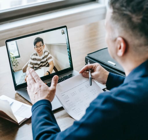Man at a desk holding a calendar page and talking to a young person on a laptop video call in a bright office setting conveying focused collaborative planning