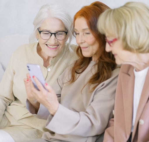 Three women in professional attire gathered closely together, each holding a mobile device and looking at the screen with warm smiles. A woman with white hair and glasses wearing a cream blazer on the left, a woman with reddish-brown hair in a beige top in the center, and a woman with blonde hair in a pink blazer on the right. The setting is a bright, neutral indoor environment. The overall tone conveys friendliness, collaboration, and engagement in a virtual meeting or group communication.