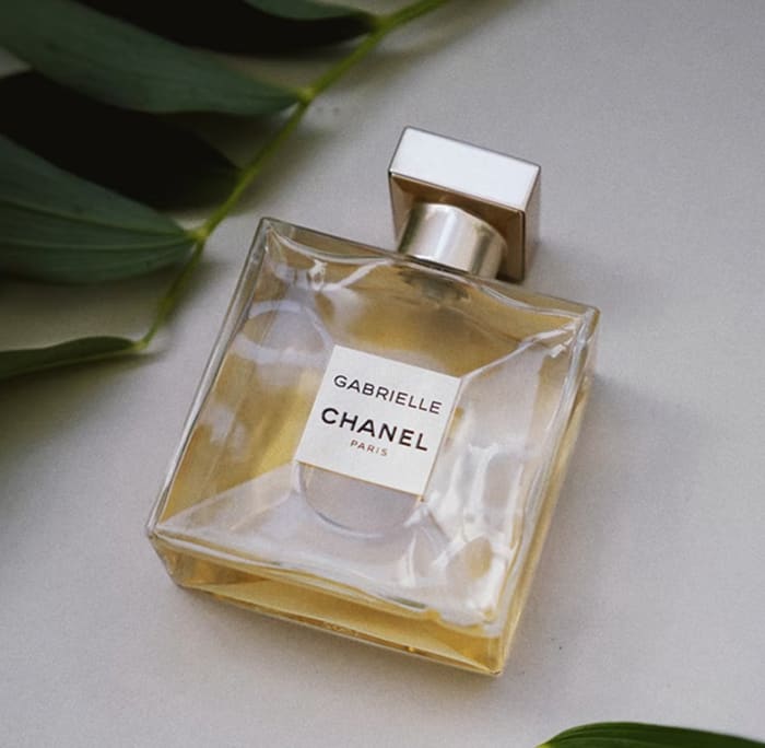 Gabrielle Chanel perfume bottle with gold liquid inside, displayed on a light surface with green plant leaves in the background, conveying luxury and elegance