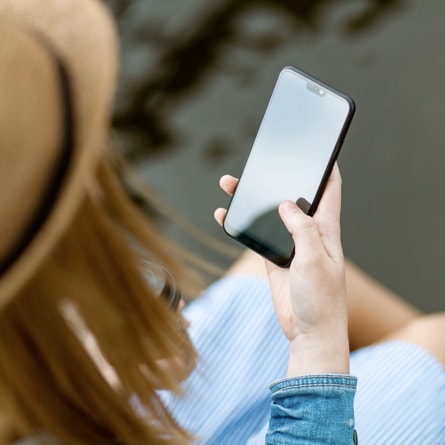 Person holding a smartphone with a blank screen, wearing a light blue striped shirt and tan hat, standing outdoors against a blurred natural background. The scene conveys a contemporary, connected lifestyle with a calm and focused mood.