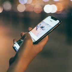 A hand holding a smartphone with a thumb interacting with the touchscreen, showing a mobile app; blurred city lights in the background at night create a warm bokeh atmosphere, conveying a calm connected mood