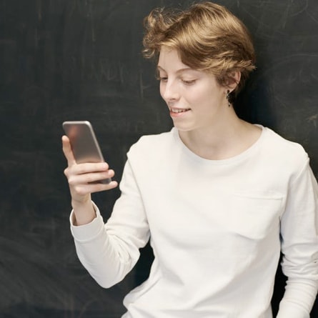 Young person smiling slightly while looking at a smartphone, standing against a dark chalkboard background; focused and content mood; no visible text in the image